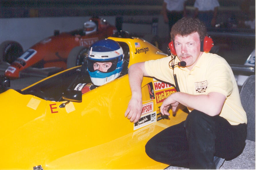 Steve in Hooters crew gear with a headset, kneeling beside the yellow Formula 2 car with driver Jernigan in the cockpit